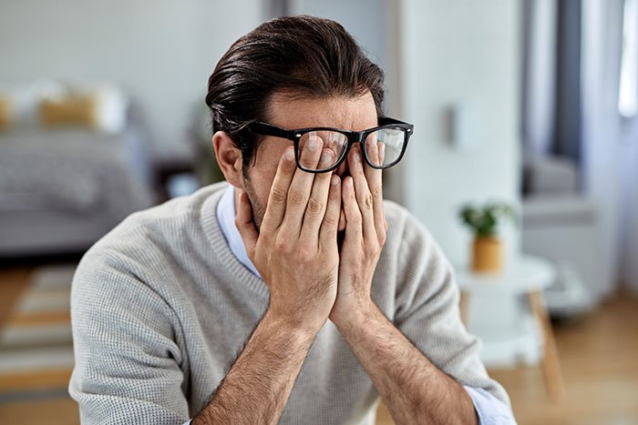 Man in glasses and gray sweater covering his face with hands, showing distress after wife&rsquo;s secret exposed.