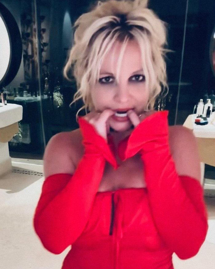 Britney Spears in a red crop top revealing bra while dancing indoors with tousled blonde hair and smoky eye makeup.