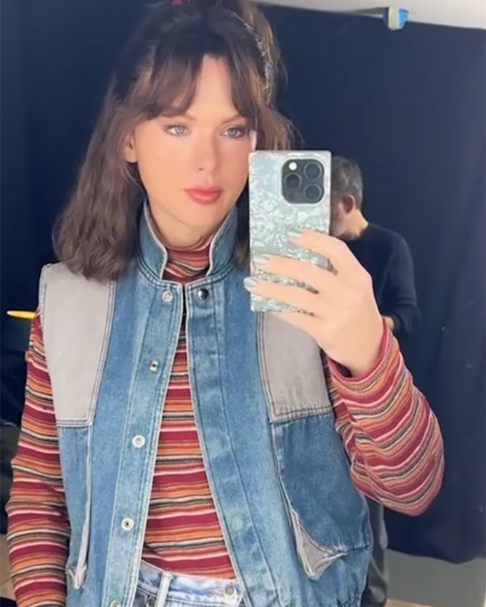 Taylor Swift taking a makeup-free selfie wearing a striped shirt and denim vest in a behind-the-scenes setting.