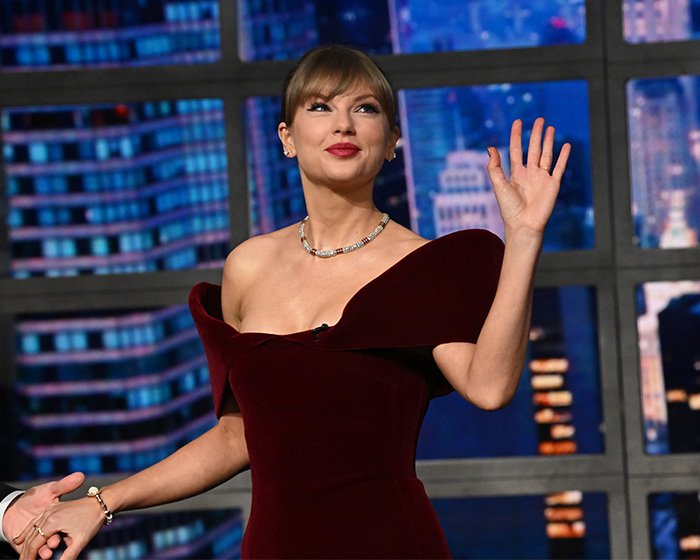 Taylor Swift smiling and waving on stage wearing a burgundy dress, highlighting her makeup-free behind-the-scenes look.