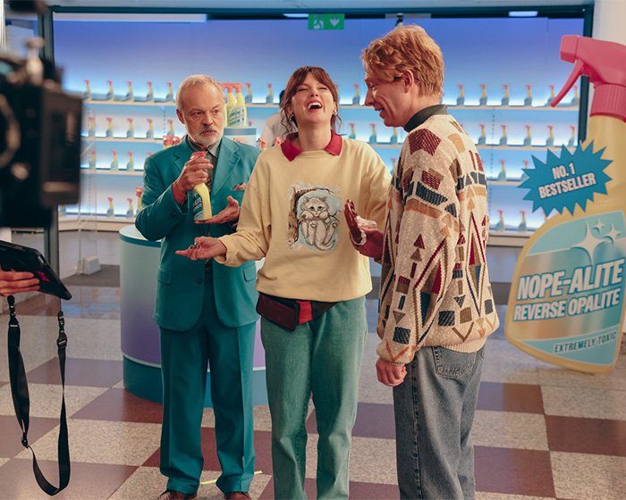 Three people laughing on a colorful set while filming a makeup-free behind-the-scenes video, capturing natural reactions.