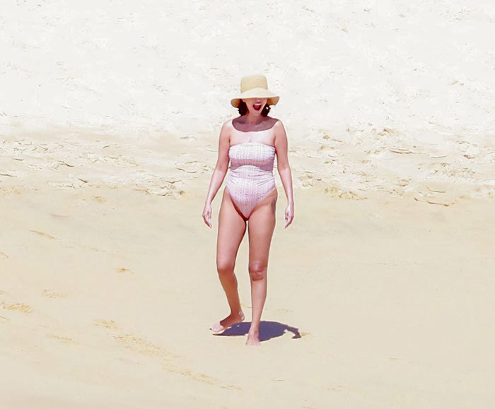 Woman in a pink swimsuit and wide-brimmed hat walking on a sandy beach, highlighting Selena Gomez swimsuit snaps debate.