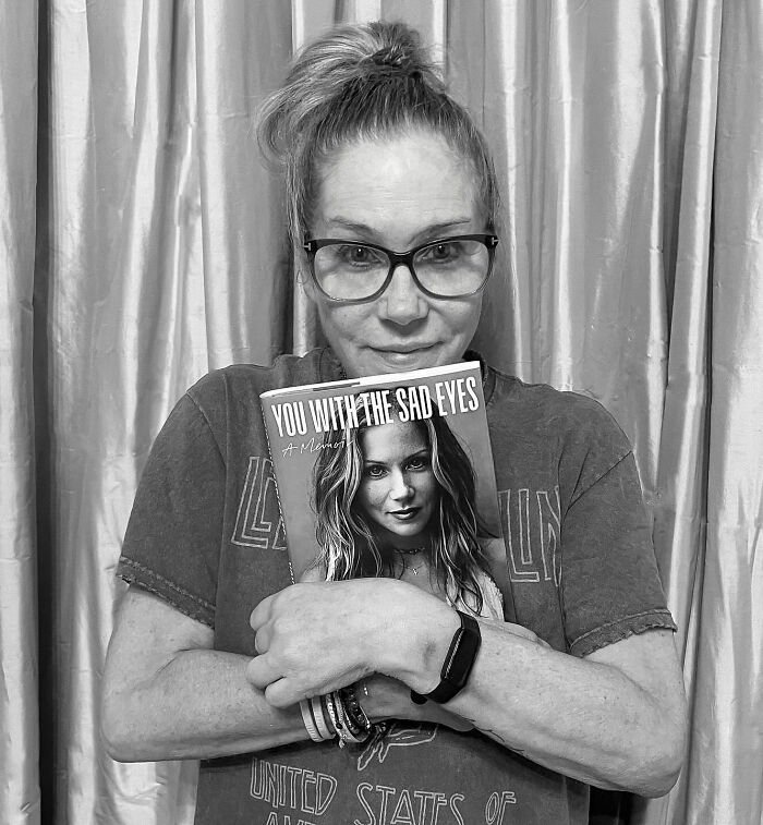 Christina Applegate wearing glasses, holding a book, sharing an update on her battle with multiple sclerosis.