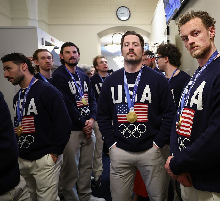Team USA men's hockey players wearing USA sweaters and gold medals, standing indoors with serious expressions.