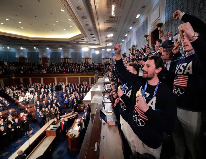 Team USA Men's Hockey players wearing Olympic sweaters, cheering passionately during a formal event at the White House.