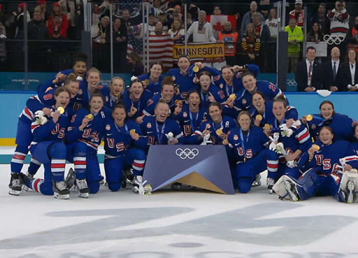 Team USA men's hockey team posing with gold medals on ice after winning at the Olympics with fans in the background.