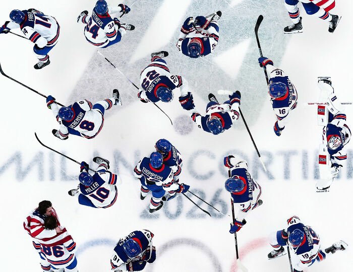Team USA men's hockey players in blue jerseys huddle on ice with sticks during a game, seen from above.