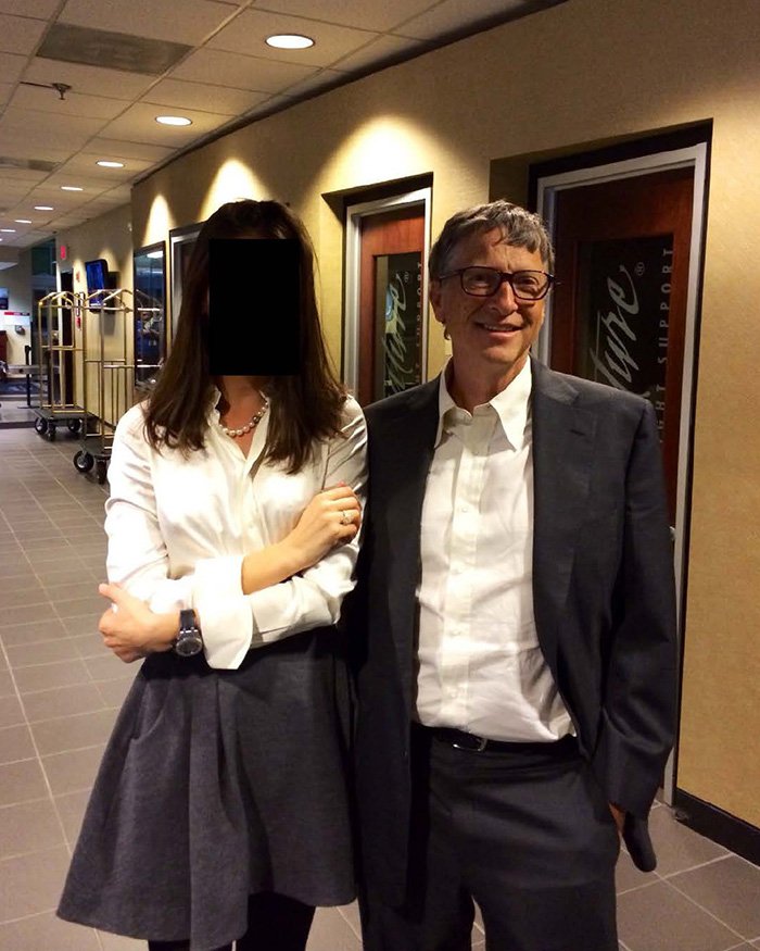 Bill Gates smiling in a suit and glasses standing next to a woman with a blurred face in a hallway setting.