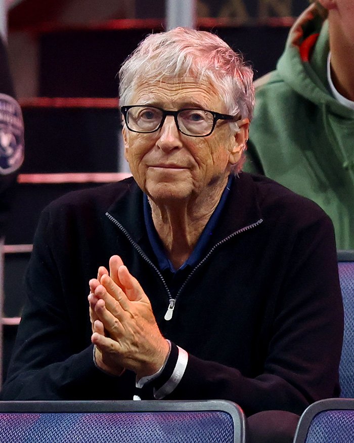 Bill Gates wearing glasses and a black zip-up sweater, seated and clasping hands amid Epstein scandal reports.