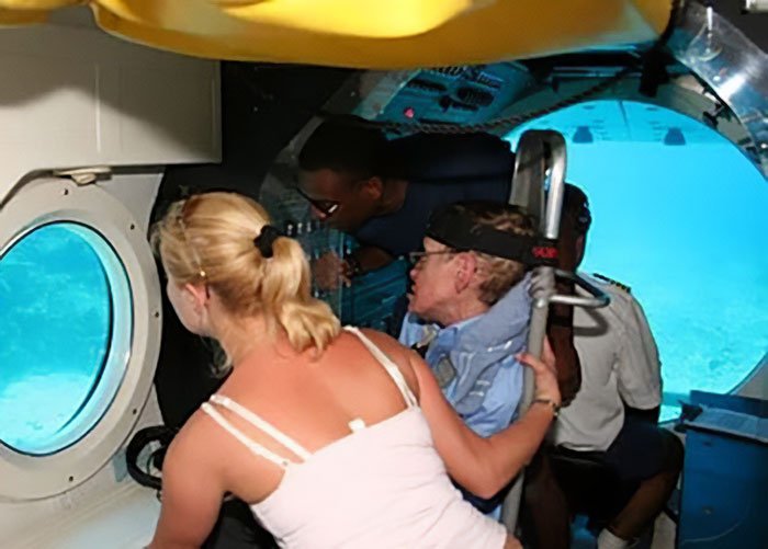 Stephen Hawking and group inside submarine, exploring underwater through round window in a confined space.