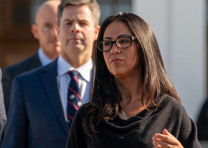 Woman in glasses and black shawl speaking outdoors during a political event about Hillary Clinton Epstein claim involving Trump.