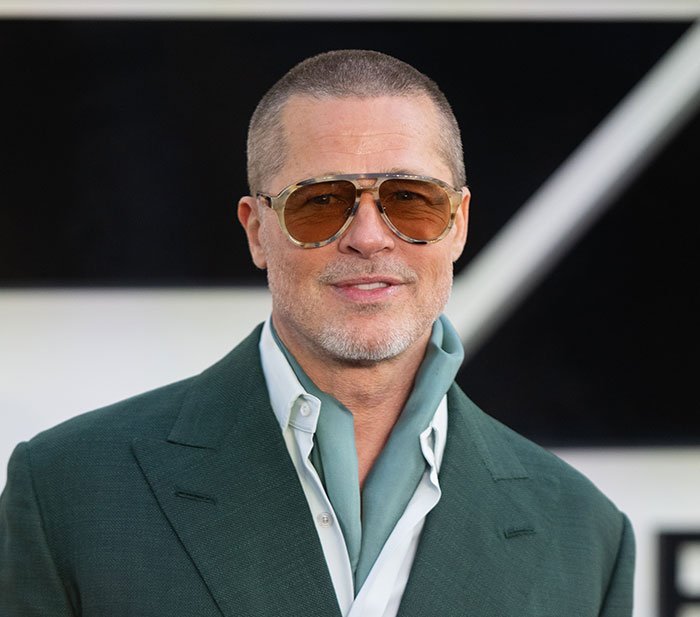 Brad Pitt wearing tinted sunglasses and green suit jacket, attending a formal event with a slight smile.