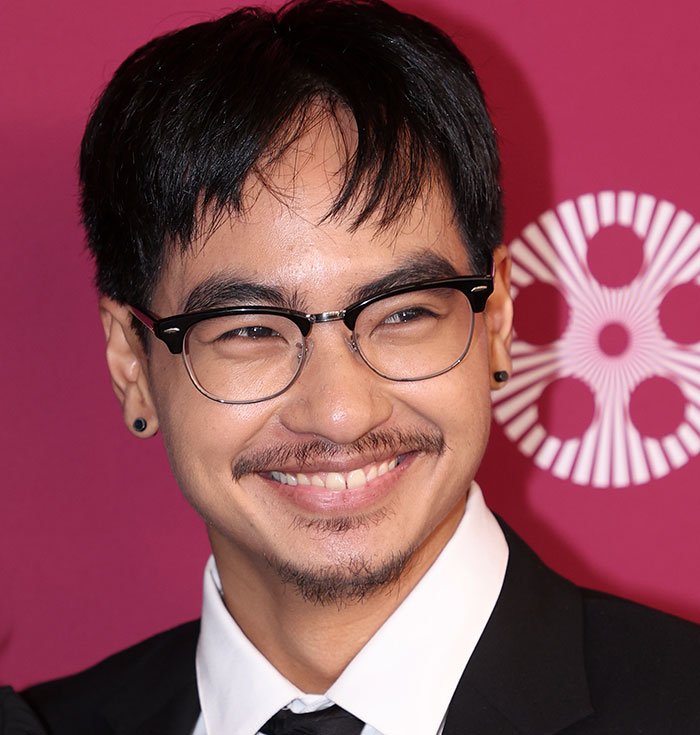 Smiling man wearing glasses and a black suit, representing Brad Pitt's kid sending a brutal message about their relationship in new movie.