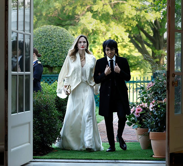Woman in white gown and person in black coat walk through garden, linked arm in arm, Brad Pitt kid relationship movie scene