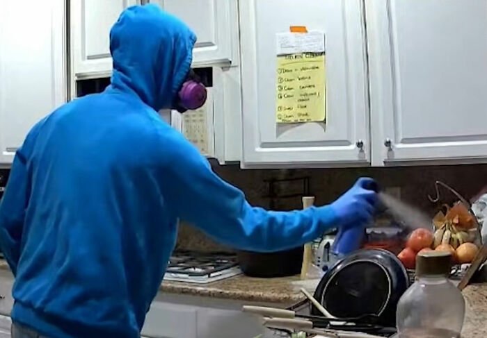 Person in a blue hoodie wearing a mask sprays disinfectant in a kitchen, linked to roommate's horrifying acts on security footage.