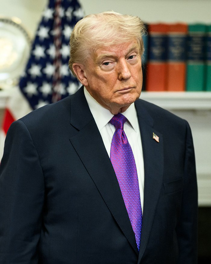 Man in a dark suit and purple tie standing indoors with books and an American flag in the background, Jeffrey Epstein’s brother discussed final conversation.