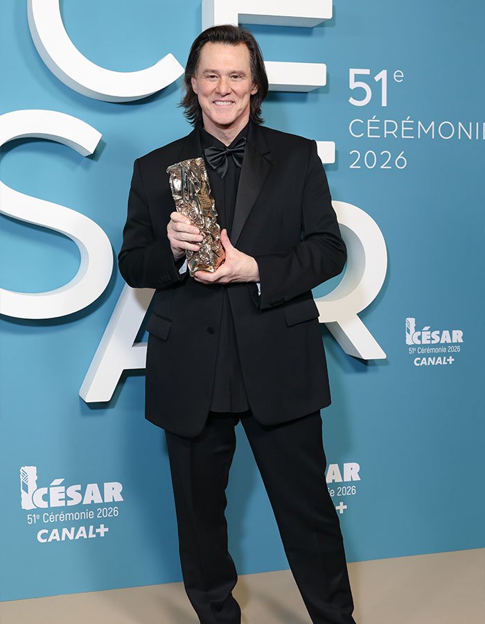 Jim Carrey holding an award, dressed in a black suit, at the 51st César Ceremony 2026 against a blue backdrop.