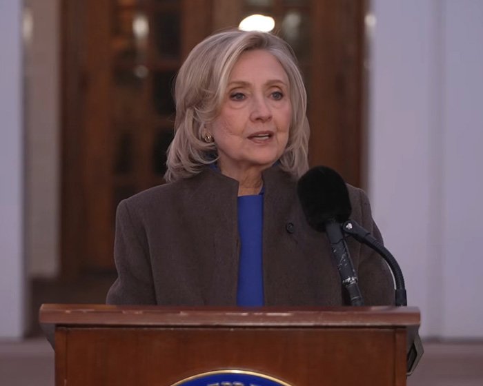 Hillary Clinton speaking at a podium, addressing Epstein ties amid Bill Clinton statement controversy.