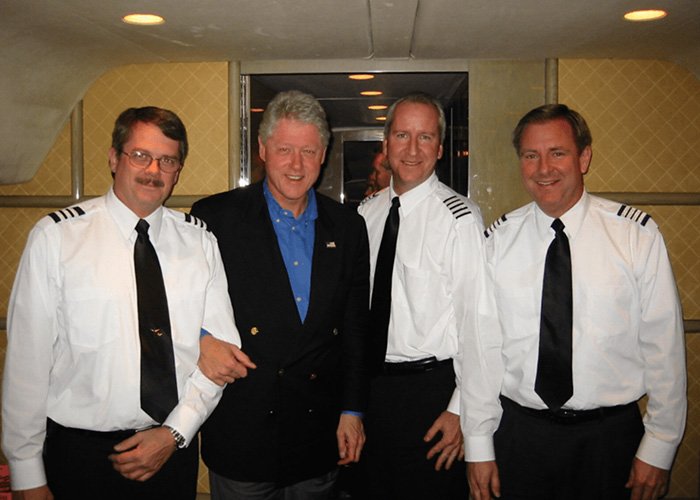 Bill Clinton posing with three men in pilot uniforms inside an airplane cabin, related to Epstein ties coverage.