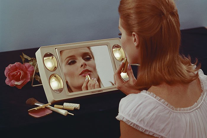 Woman applying lipstick while looking in mirror, highlighting beauty standards and their impact on women.