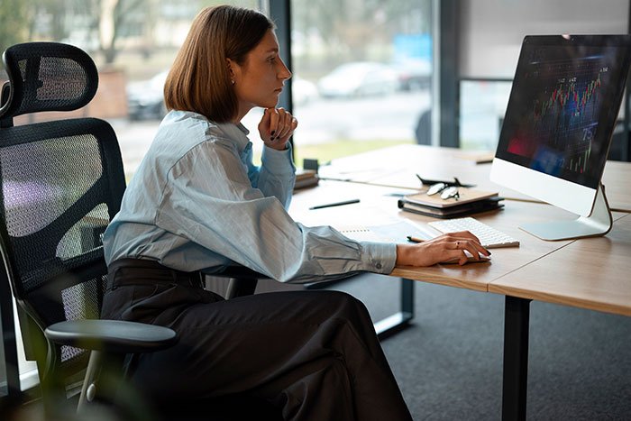 Woman sitting at desk, thoughtfully looking at computer screen, illustrating concepts of beauty standards and their impact on women.