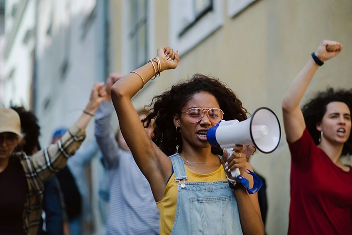 Woman wearing glasses and denim overalls uses megaphone leading a protest against beauty standards with raised fists.