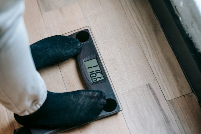 Person wearing black socks standing on digital scale showing low weight on wooden floor near window.