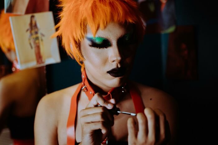 Person with vibrant orange hair and bold makeup focused on applying nail polish, related to narcissist quiz concept.