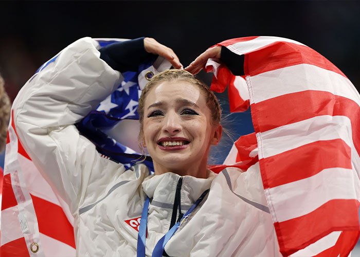Amber Glenn smiling with tears, wrapped in the American flag, reflecting the backlash and threats over her beliefs.
