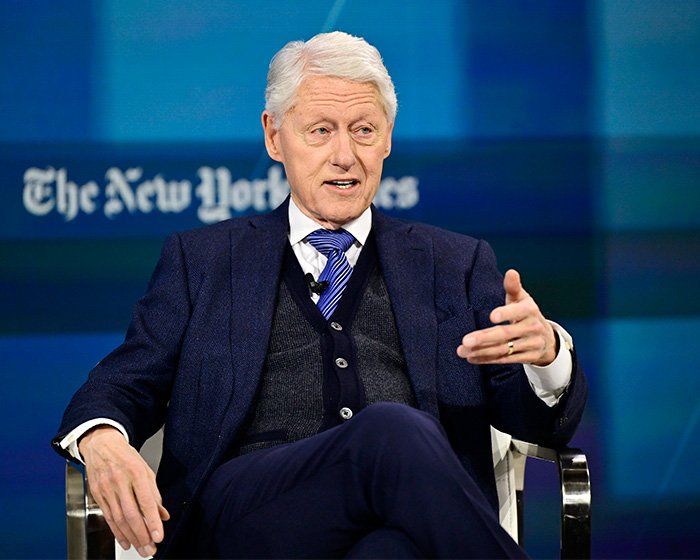 Bill Clinton seated in a suit and tie, speaking during an interview on a stage with The New York Times backdrop.