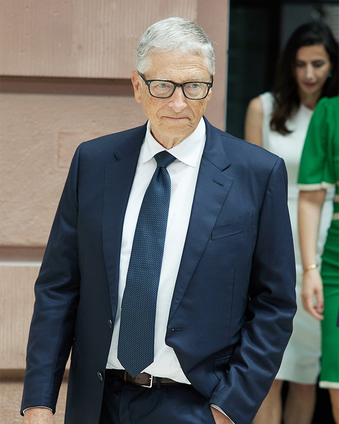 Bill Gates wearing glasses and navy suit, walking outside with people in background amid Epstein scandal news.