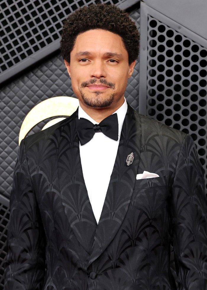 Trevor Noah wearing a patterned black tuxedo with a bow tie, posing against a geometric black and gold backdrop.
