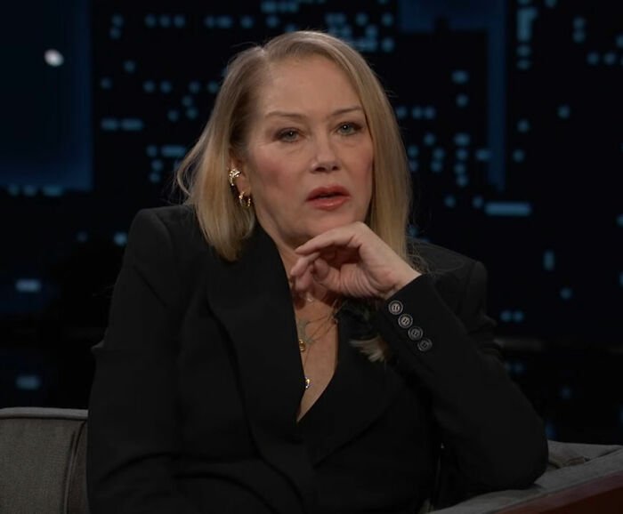 Christina Applegate in a black blazer giving an emotional update on her battle with multiple sclerosis on a talk show set.