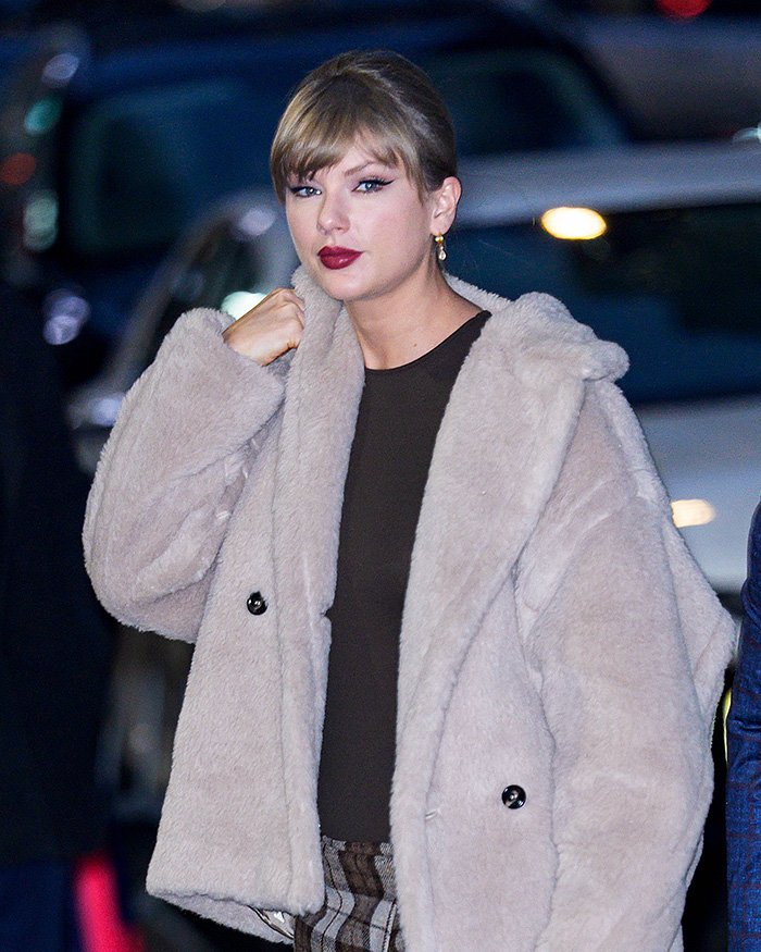 Female indie musician in a beige fur coat and dark top, posing outdoors with a confident expression at night.
