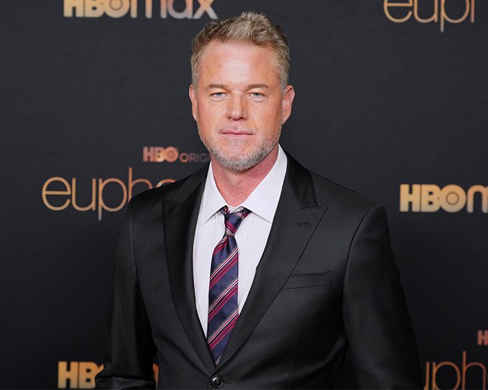 Eric Dane posing at a formal event wearing a dark suit and striped tie with HBO Max branding in the background.