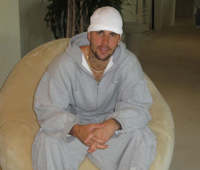 Justin Bieber wearing gray tracksuit and white cap sitting on a beige chair related to Epstein theory controversy.