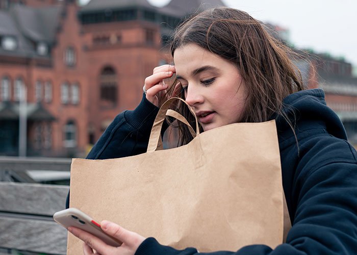 Young woman holding a paper lunch bag and looking at her phone, illustrating workplace entitlement and lunch conflict.