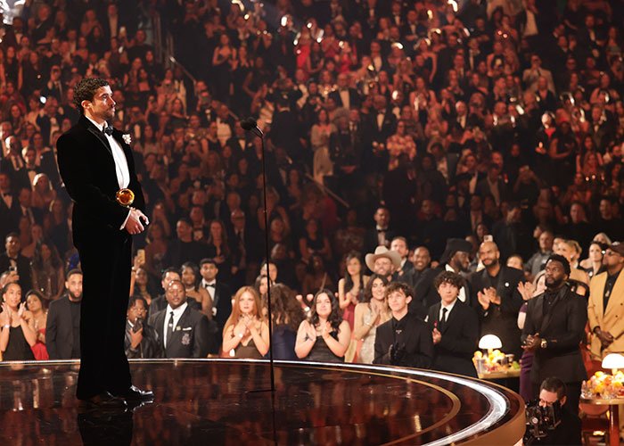 Man in tuxedo holding Grammy on stage with large audience applauding, showing Grammy seat filler event atmosphere.