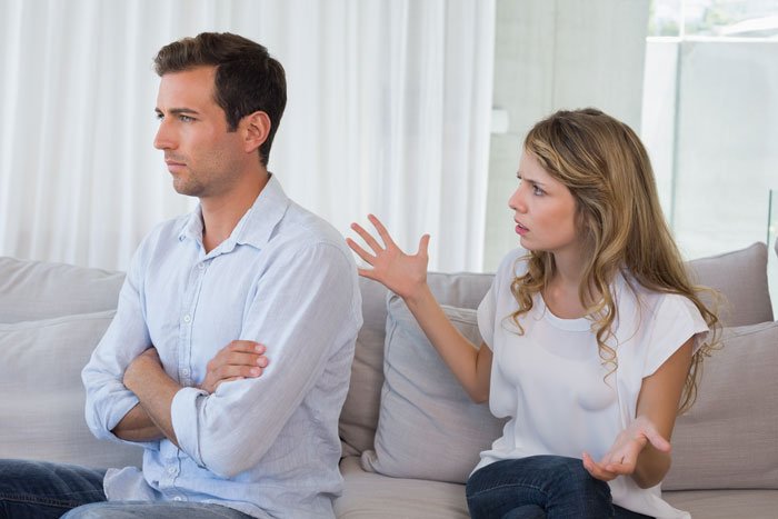 Young man and woman arguing on couch, man appears upset after discovering fiancée is keeping secrets from him.