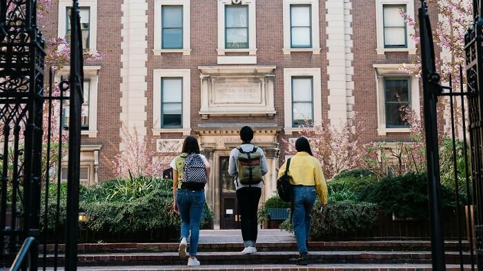 Three students with backpacks walking toward a college building, illustrating challenges of getting into college nowadays.