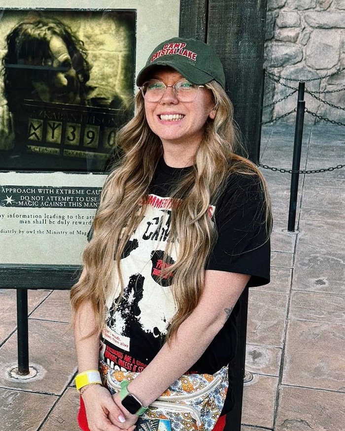 Young woman smiling outdoors, wearing glasses and a cap, with long hair and casual clothing, symbolizing danger and escape awareness.