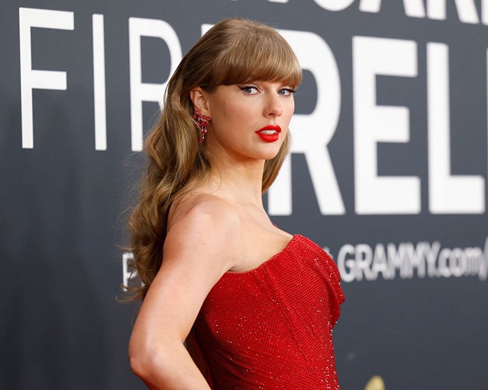 Taylor Swift posing at an event in a red dress with red lipstick, related to makeup-free behind-the-scenes video reactions.