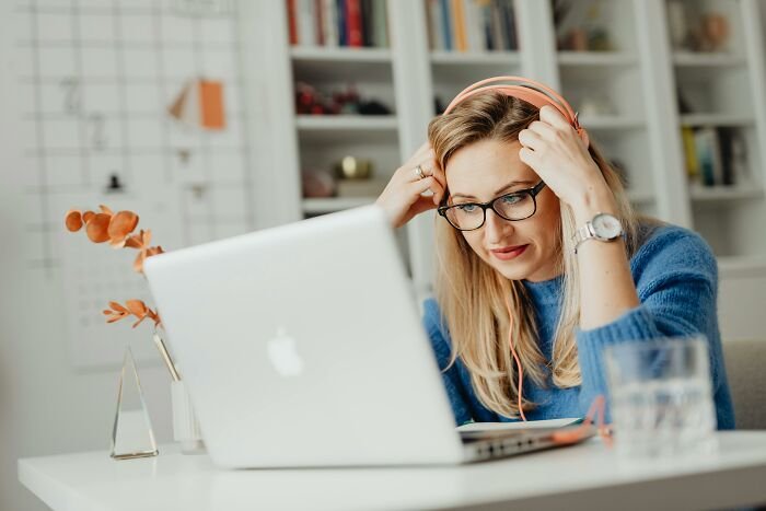 Woman wearing glasses and headphones focused on solving a rebus quiz on a laptop at a home office.