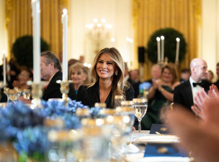 Melania Trump smiling at governors dinner event, wearing a black outfit amid formal table setting and guests applauding