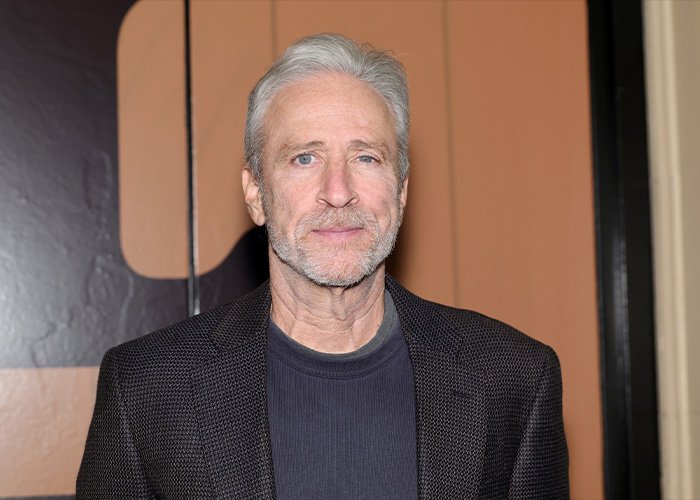Jon Stewart in a blazer and dark shirt looking at the camera during an indoor event related to Epstein files news.