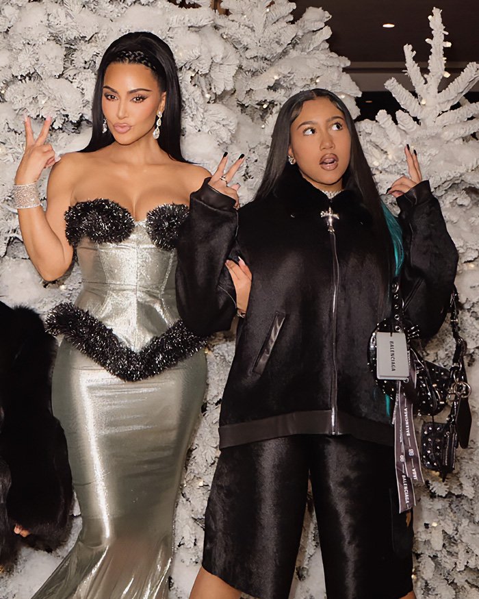 Kim Kardashian and a woman posing with peace signs at an event with white winter-themed decor behind them.