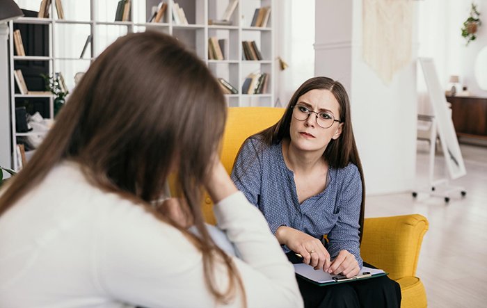Woman sitting in a yellow chair listening intently to another woman, highlighting themes of submissive nature and standing up for herself.