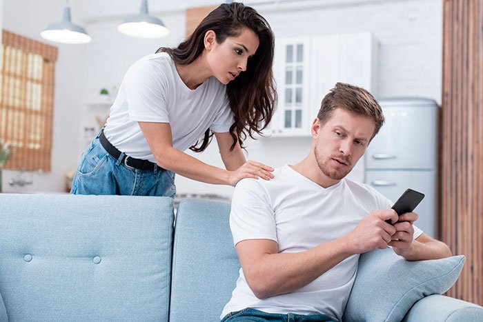 Married man texting woman he loved years ago, wife looking concerned while he holds a phone on couch at home.