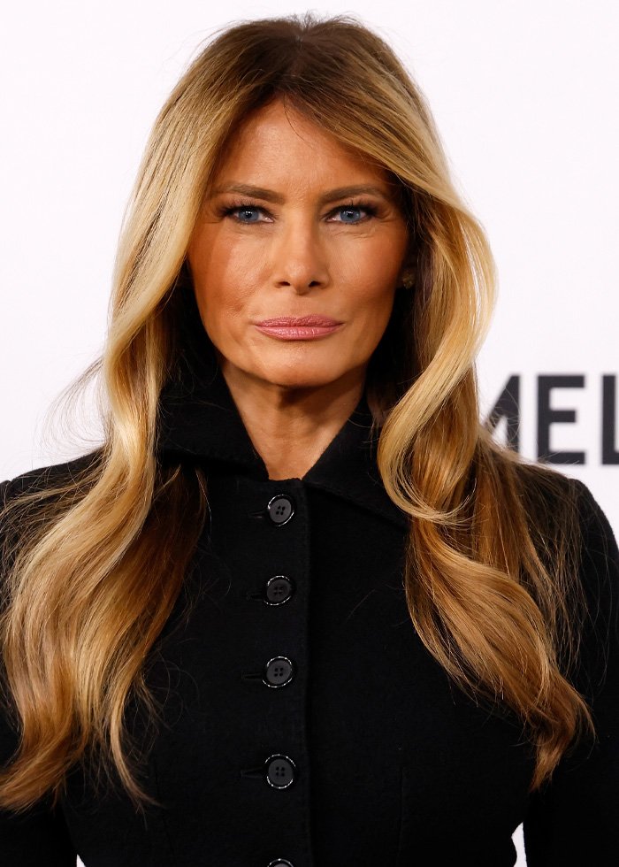 Melania Trump with long blonde hair and polished makeup wearing a black coat in a formal setting