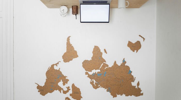 Cork board with creatively shaped world map pieces mounted on a white wall under a digital clock and laptop screen.